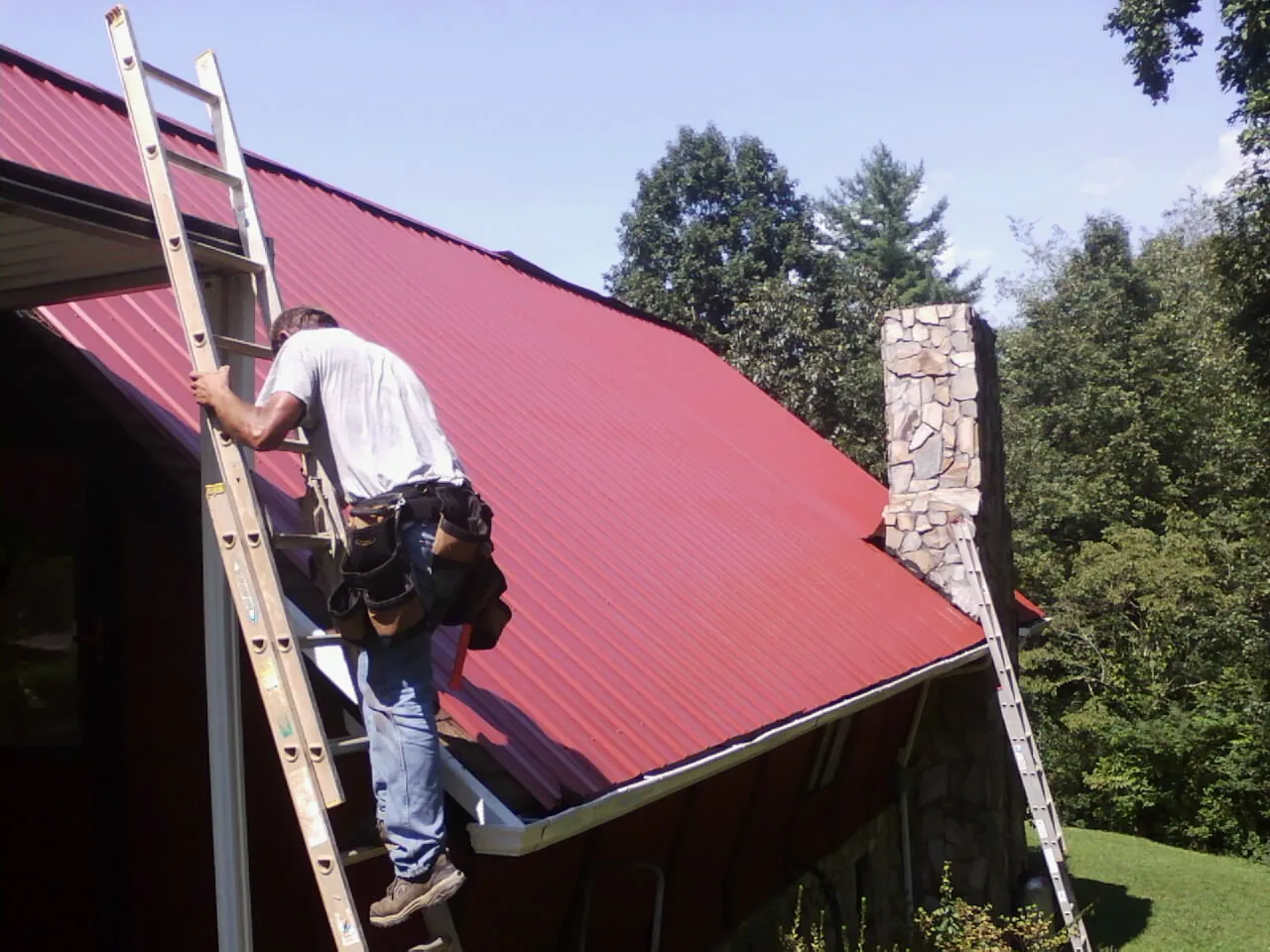 Roofer on a ladder beside a red metal roof and stone chimney — Soffit & Fascia Repair by Roofing Brothers in Holmen